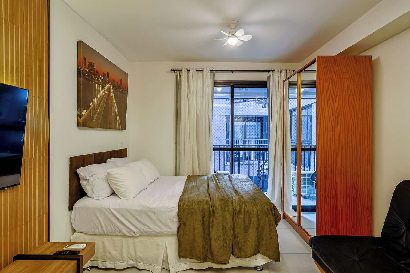 A hotel room with a bed, a TV, a painting, a balcony with curtains, and a ceiling fan.