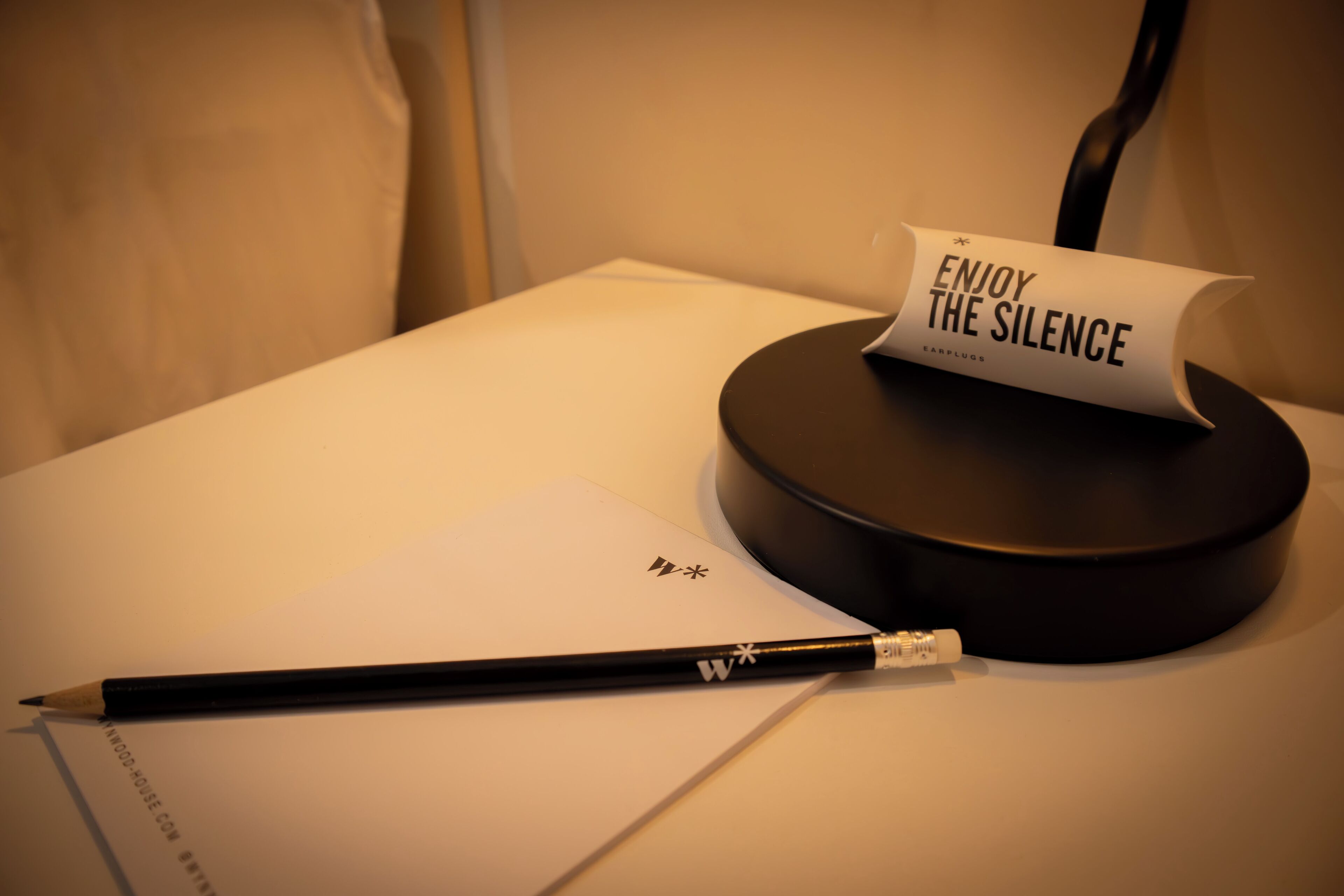 Una taula amb una làmpada de sobretaula negra, un llapis negre, un tros de paper amb un logotip i un cartell que diu "ENJOY THE SILENCE".