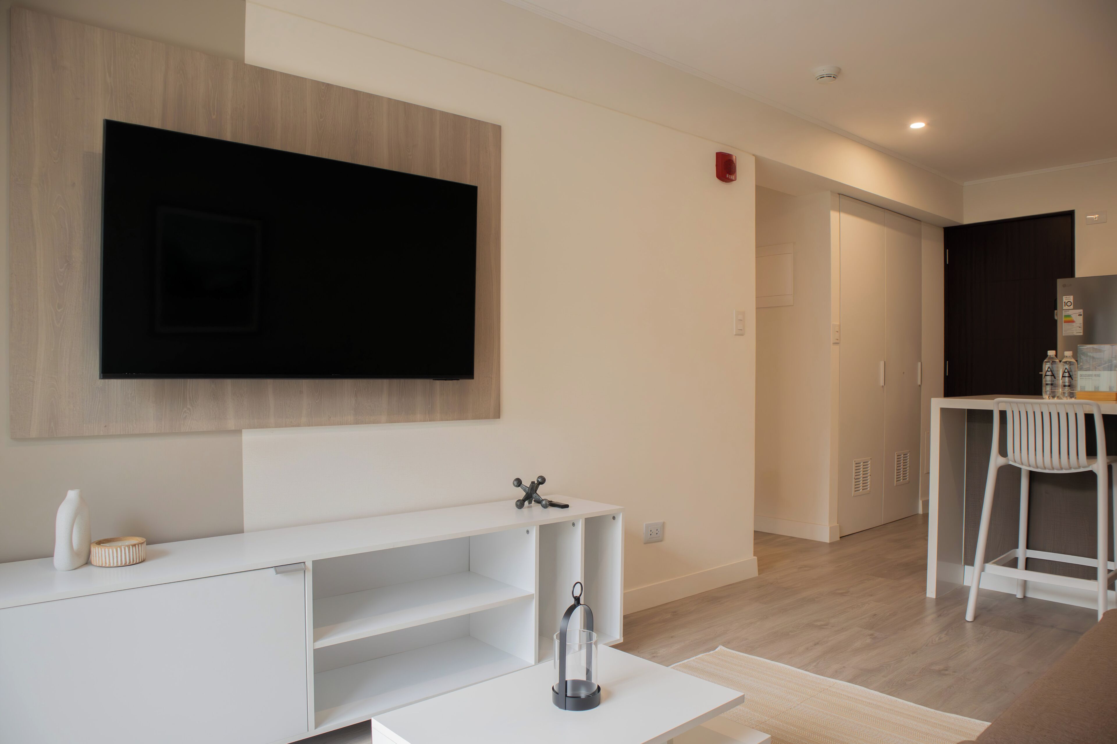 Una sala de estar moderna con un televisor de pantalla plana montado en la pared, un mueble blanco para entretenimiento y una pequeña mesa con un recipiente de vidrio.