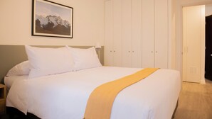 Ein ordentlich bezogenes Bett mit weißen Laken und einer gelben Tagesdecke, ein gerahmtes Bild von Bergen an der Wand und ein geschlossener Kleiderschrank.
