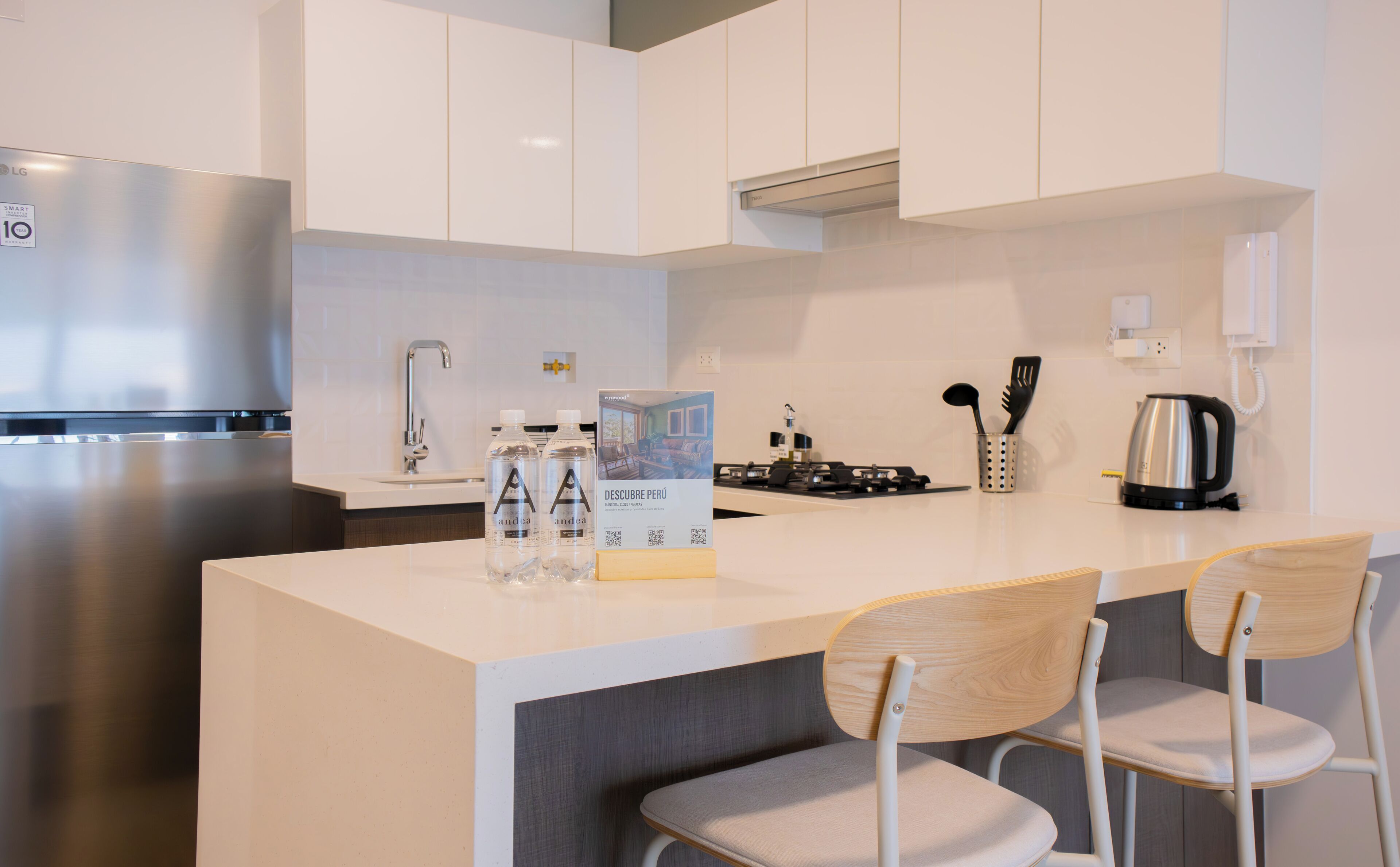 A modern kitchen with a stainless steel refrigerator, a gas stove, and a coffee maker. There are two chairs with wooden seats and metal frames.