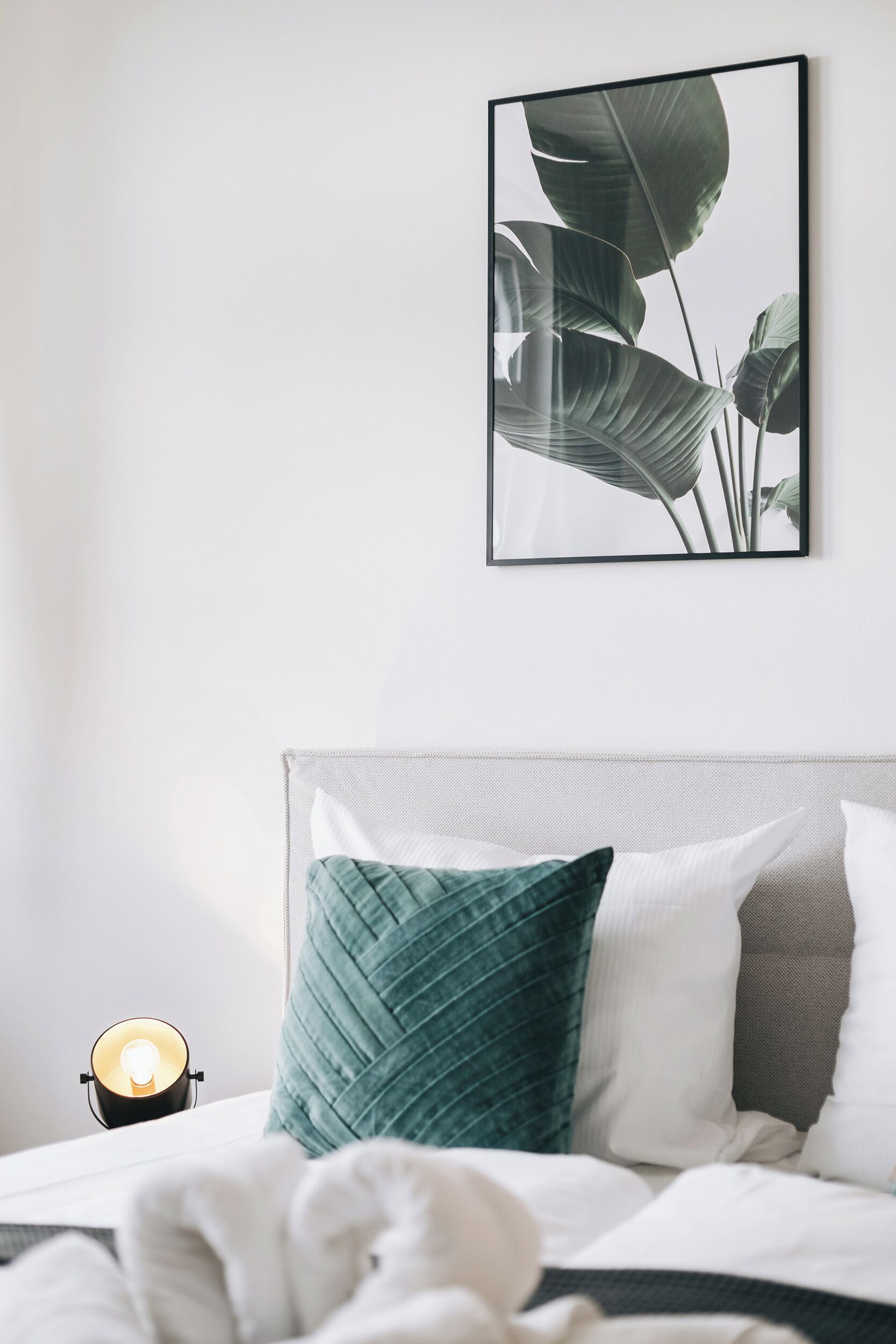 Un lit bien fait, orné d’un coussin vert, d’un coussin blanc et d’un coussin gris. Au-dessus du lit, un tableau encadré représente des feuilles.