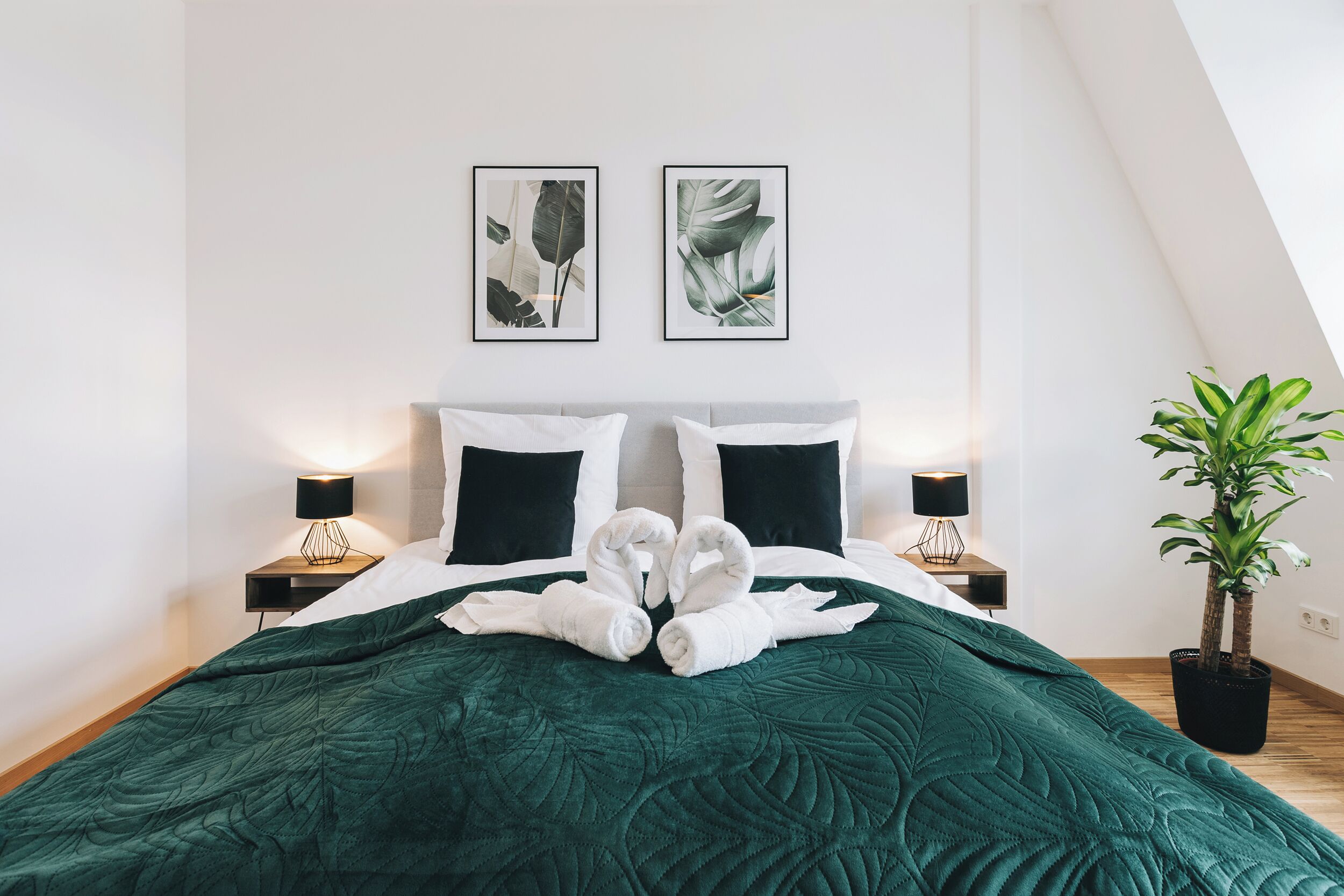 Un lit bien fait, recouvert d’une couette verte, deux gravures encadrées représentant des motifs botaniques accrochées au mur, et une plante en pot placée à côté du lit.