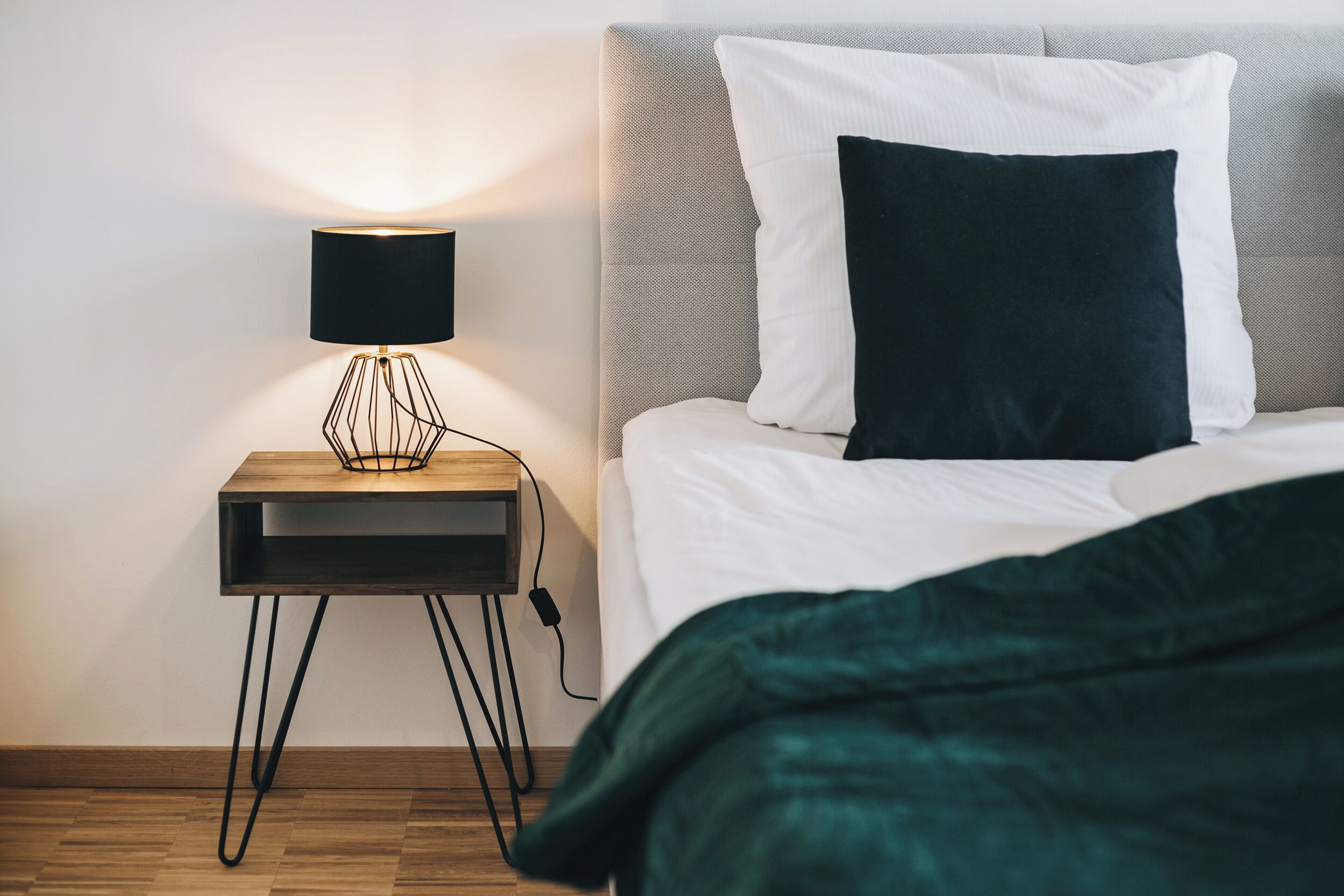 A neatly made bed with white and dark green bedding, a bedside lamp, and a small wooden nightstand with a black lamp.