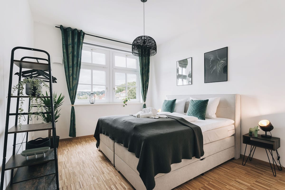 A bedroom with a bed, a window with curtains, a bedside table with a lamp, and a wall-mounted shelf with plants.
