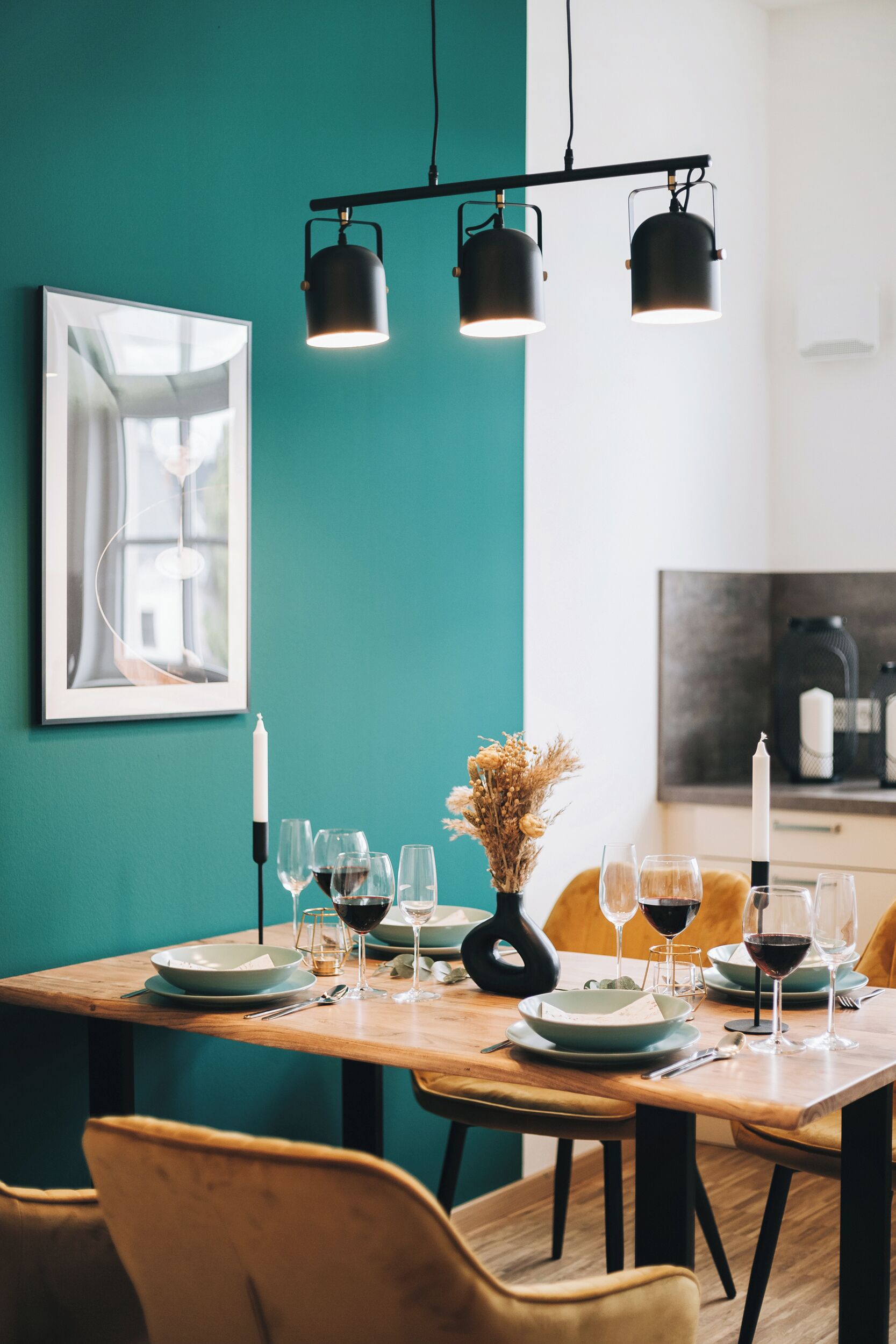 A dining table set with plates, glasses, and a candle, with teal walls and a modern pendant light fixture.