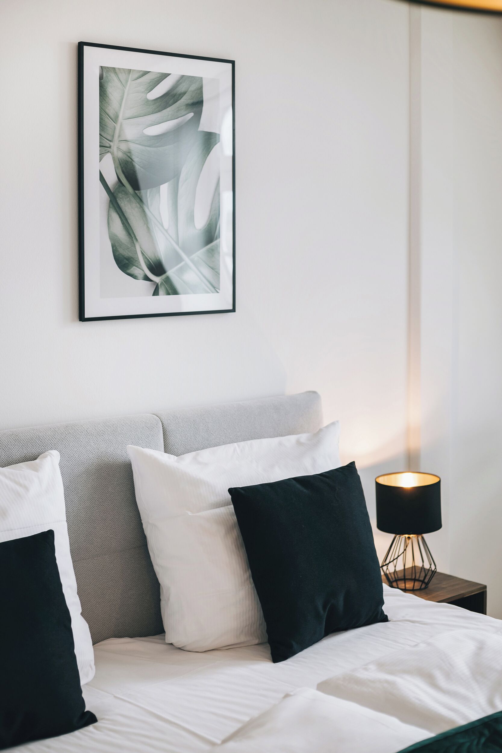 Ein ordentlich bezogenes Bett mit weißer und schwarzer Bettwäsche, ein gerahmtes Pflanzenbild an der Wand und eine Nachttischlampe.