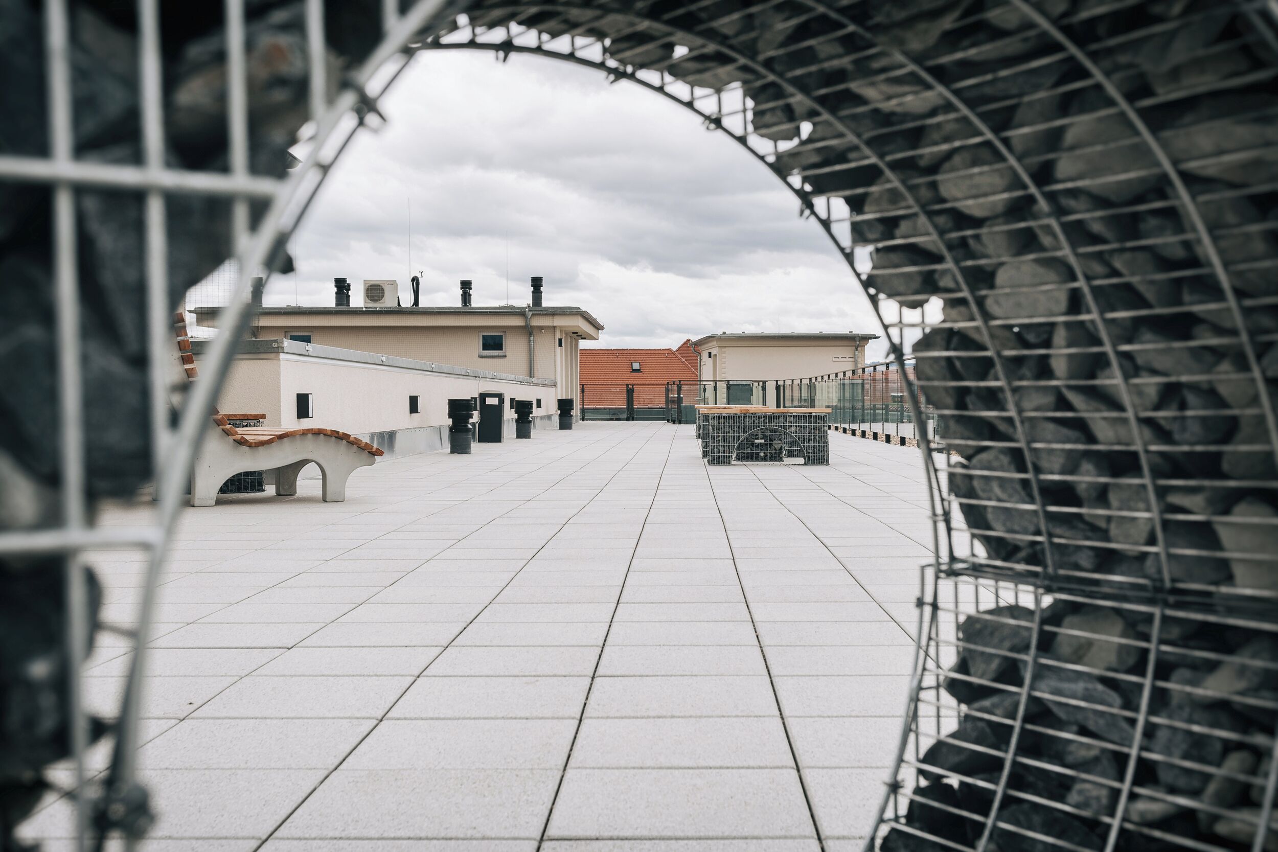 A tiled rooftop with a metal railing, buildings in the background, and a cloudy sky.