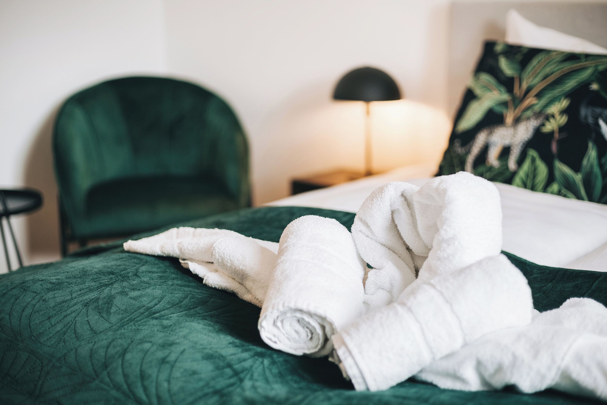 A neatly made bed with a green quilt, a rolled white towel, a black lamp, and a green chair in a hotel room.