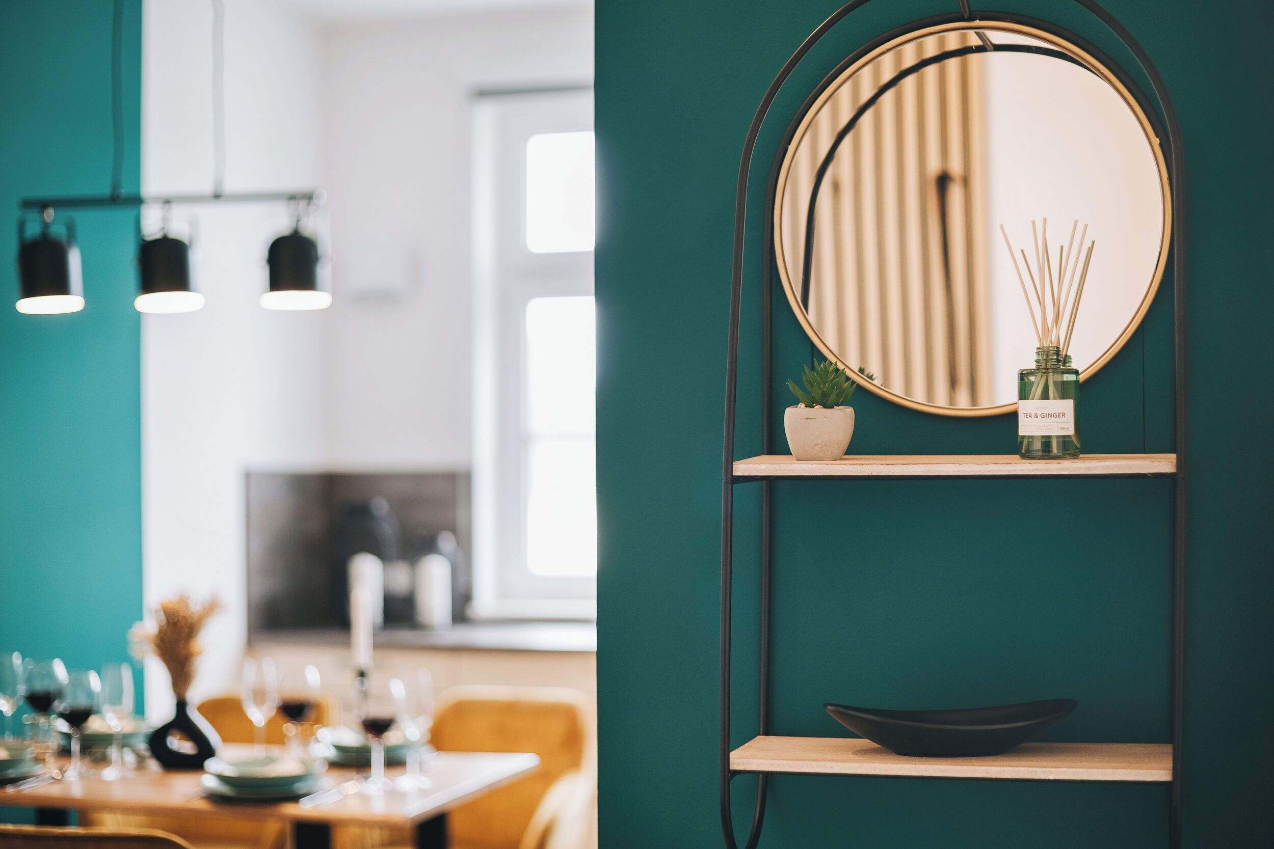 Ein Essbereich mit einer türkisfarbenen Wand, einem runden Spiegel mit Holzrahmen und einem kleinen Regal mit Pflanze und Reed-Diffuser.