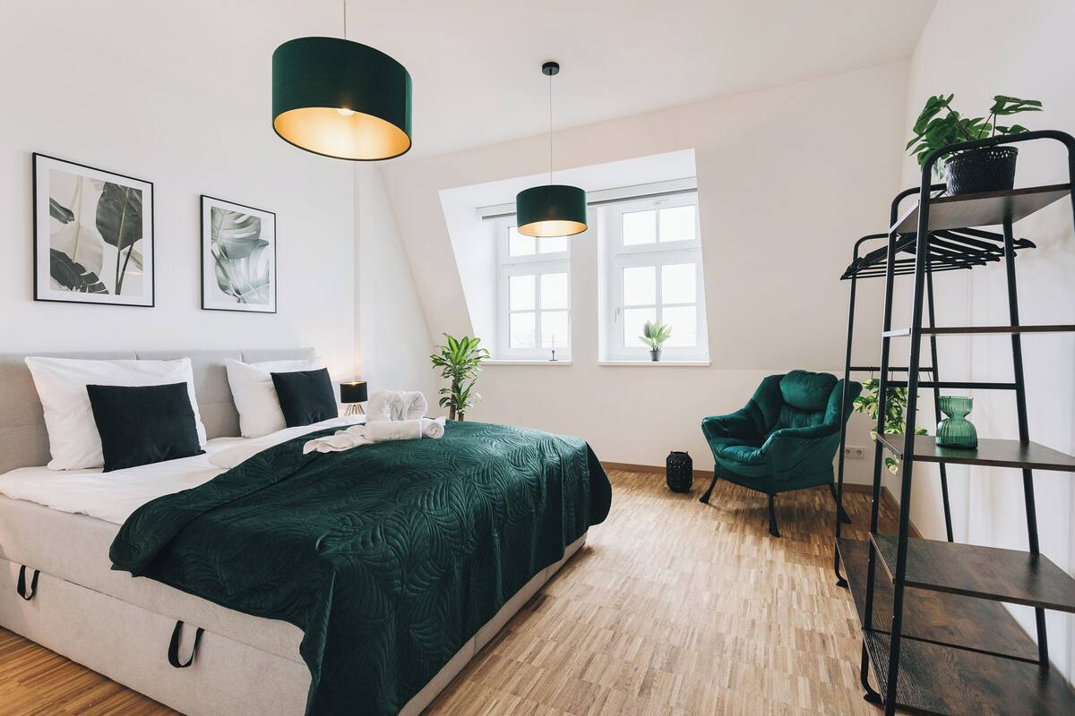 A modern bedroom with a bed, two pendant lights, a green chair, a black metal shelf, and two windows.