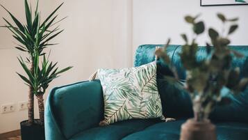 Un sofá azul verdoso con un cojín estampado con hojas, una planta alta en maceta y un cuadro en la pared.