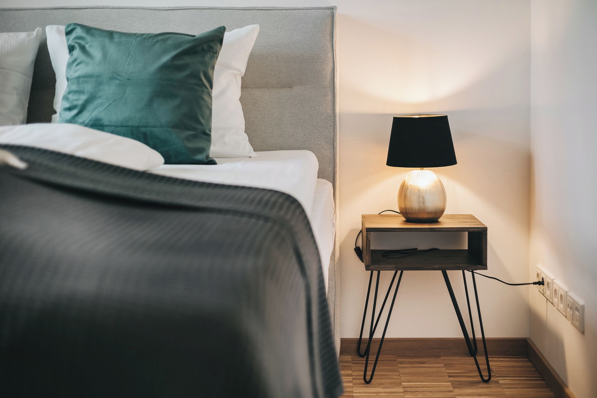 A neatly made bed with a dark blanket, a teal pillow, and a white pillow. A small wooden nightstand with a black lamp and a power outlet is beside the bed.
