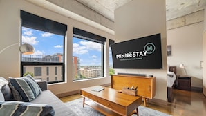 A modern living room with a large TV displaying 'Minnestay Premier Vacation Homes', a sofa, a coffee table, and large windows offering a city view. - Sable 603 - Alcove (Minneapolis)