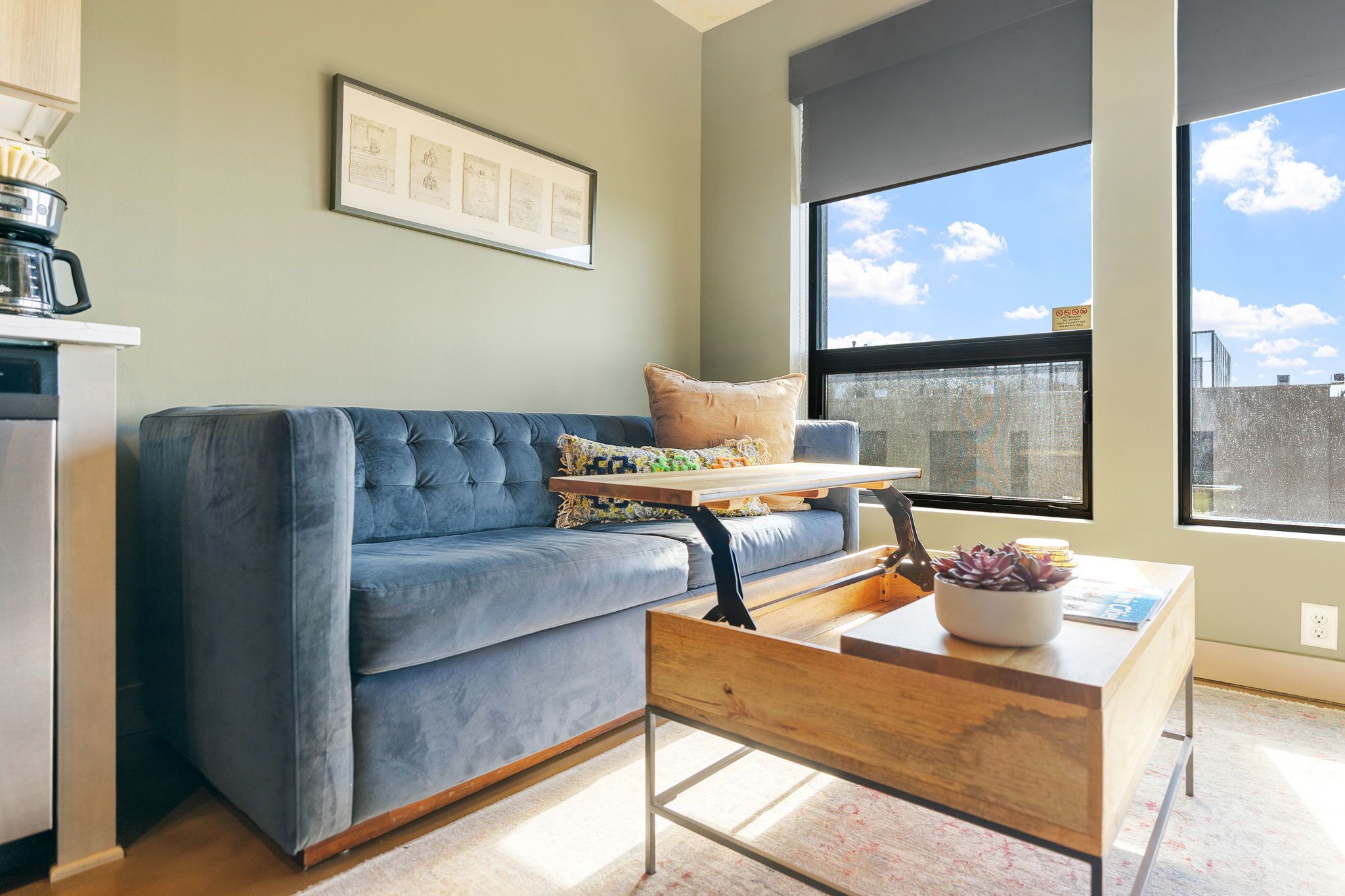 A modern living room with a blue sofa, a wooden coffee table, and a large window offering a city view.