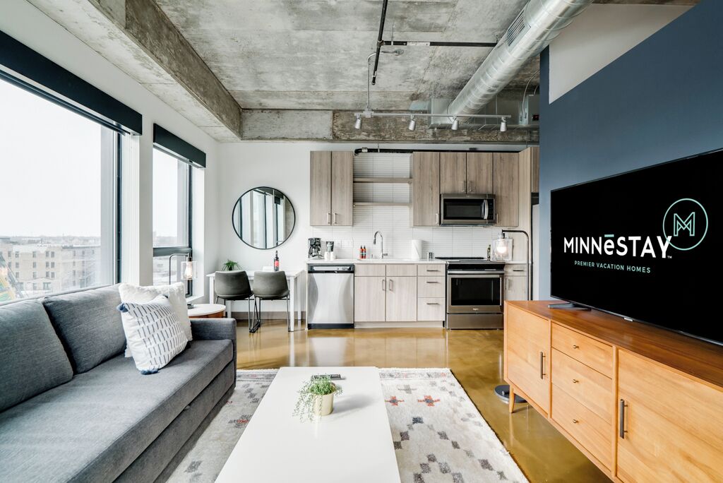 Una sala de estar moderna con un sofá gris, una mesa de centro con una planta, un televisor de pantalla plana y una zona de cocina con armarios de madera y un microondas.
