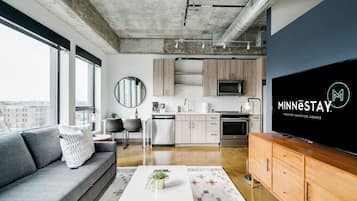 Una sala de estar moderna con un sofá gris, una mesa de centro con una planta, un televisor de pantalla plana y una zona de cocina con armarios de madera y un microondas.
