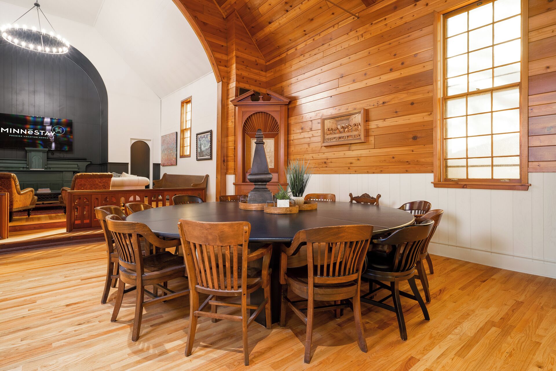 A wooden-paneled dining room with a large round table and chairs, a chandelier, and a fireplace.