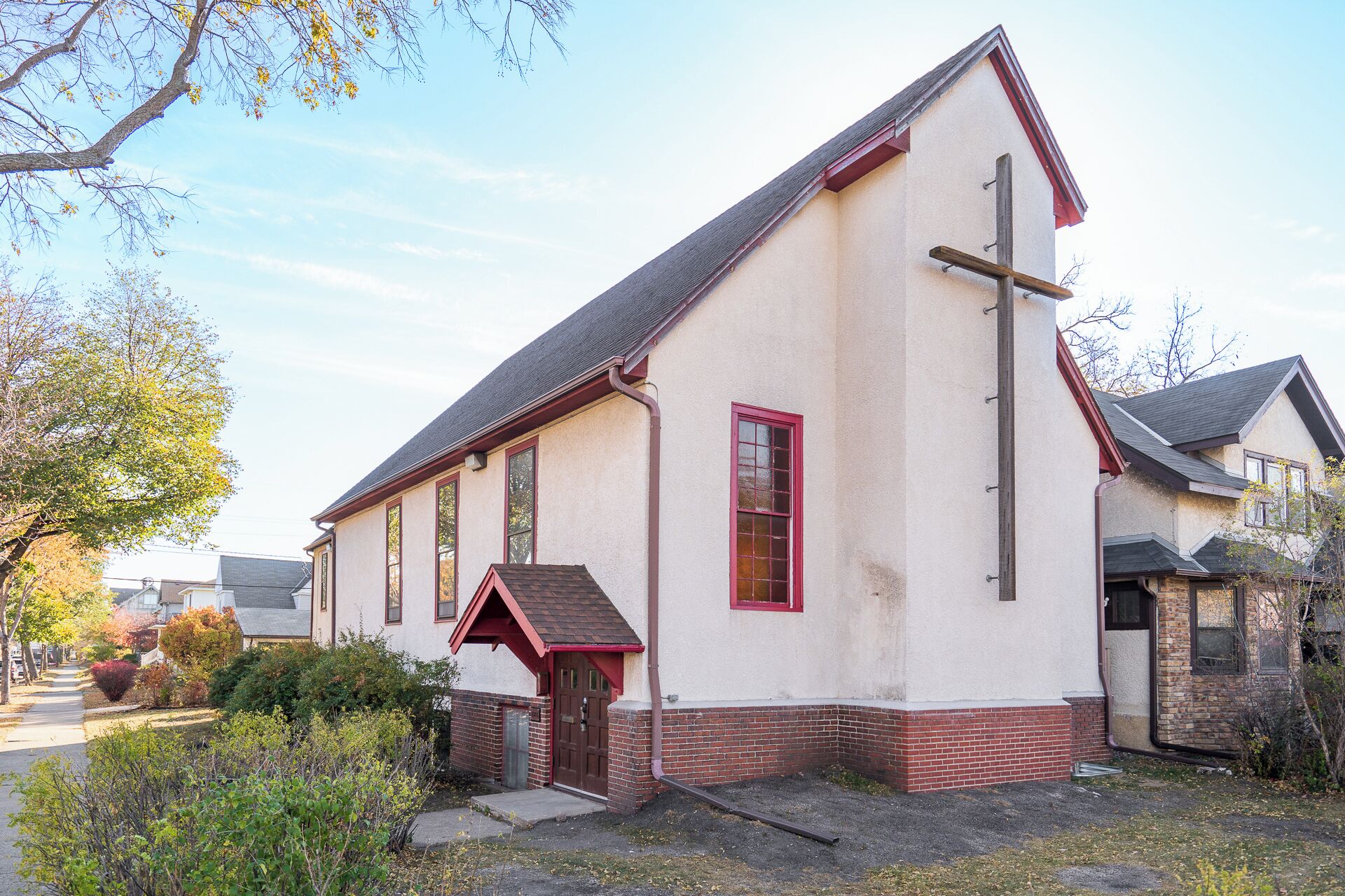A small church with a steep gable roof, a wooden cross, and a brick foundation.