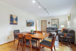 A modern living room with a dining area, featuring a wooden dining table, grey sofas, and a large window with a view of the outdoors. - Cherokee Park Family Escape (St. Paul)