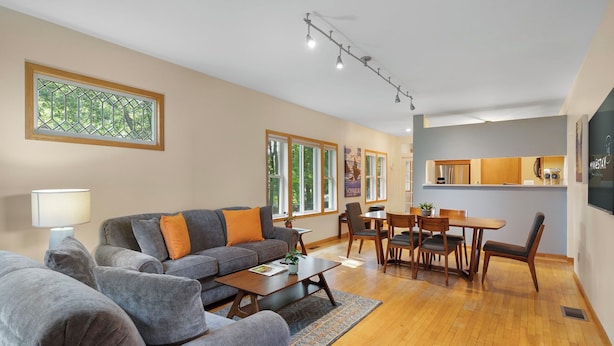 A modern living room with a sofa, coffee table, dining table, and chairs. There is a window with a view of trees, a kitchen area, and a lamp. - Cherokee Park Family Escape (St. Paul)
