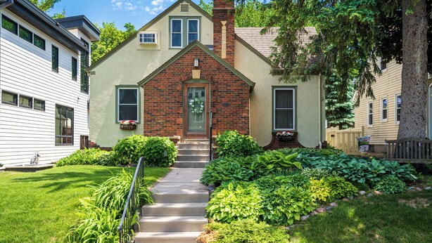 A two-story house with a brick chimney, a front door with a wreath, and a well-maintained garden with various plants and a pathway leading to the door. - Edina Escape (Minneapolis)
