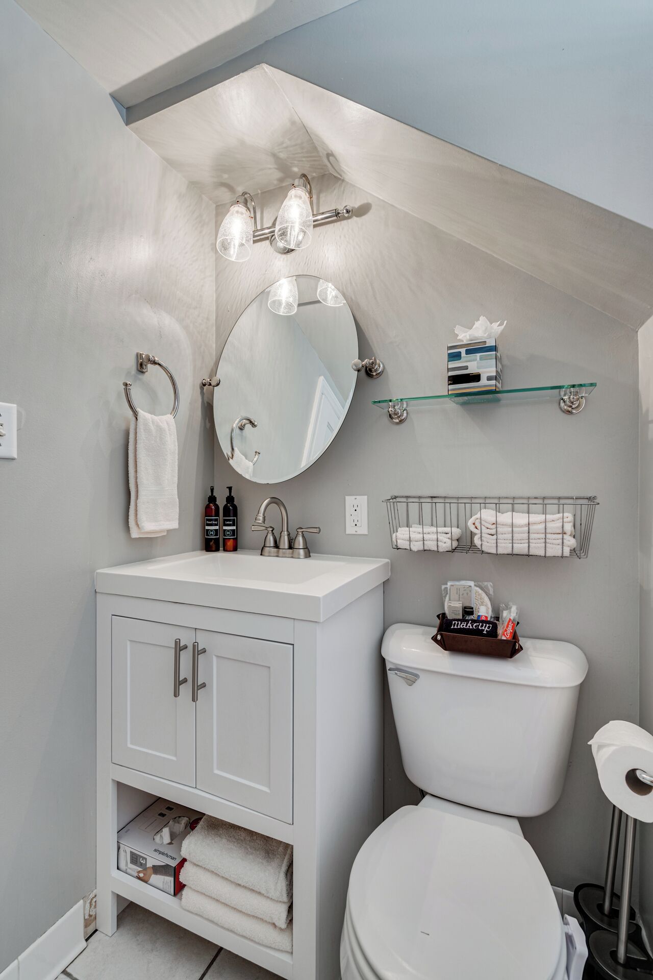 A modern bathroom with a white vanity, a round mirror, a toilet, and a small shelf with toiletries.