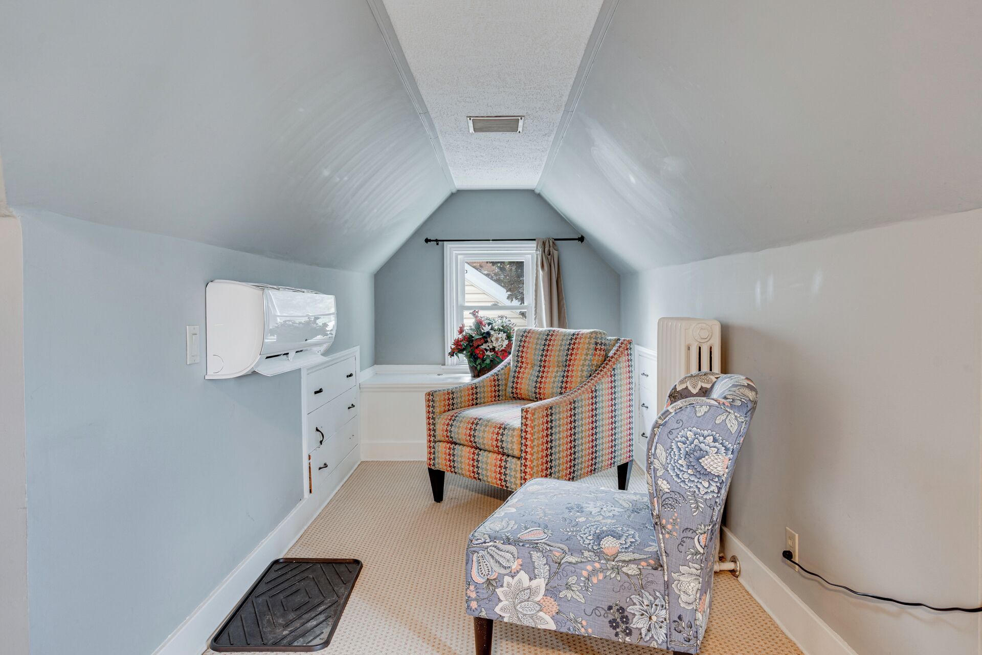 A narrow attic room with a sloped ceiling, a window with curtains, a white radiator, a patterned armchair, and a small table with a vase of flowers.