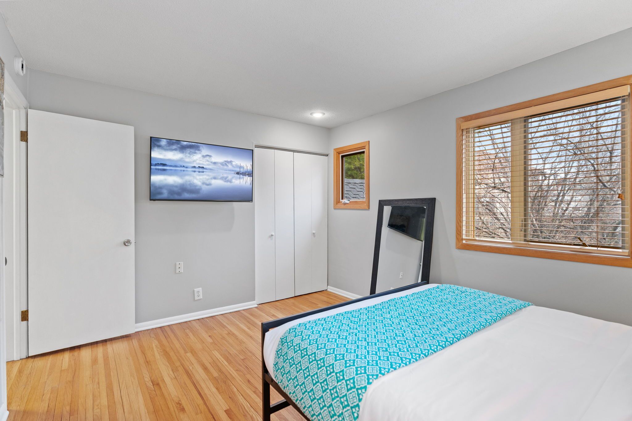 Ein Schlafzimmer mit Bett, Fenster mit Jalousien, Tür und einem an der Wand befestigten Fernseher.