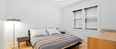 Une chambre à coucher comprenant un lit, des tables de chevet, une lampe sur pied et une fenêtre avec des stores.