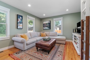 A living room with a sectional sofa, a brown leather ottoman, a patterned rug, and artwork on the walls.