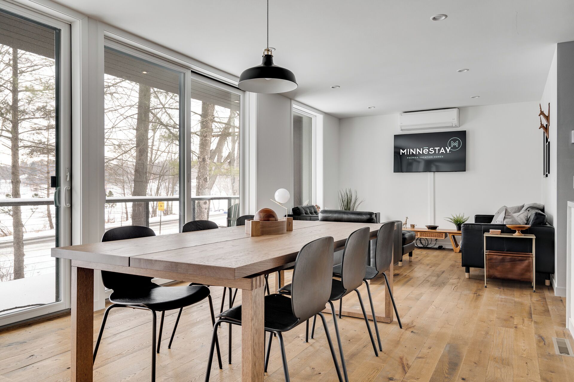 A modern living room with a large TV, a dining area with a wooden table and chairs, and a view through large windows.