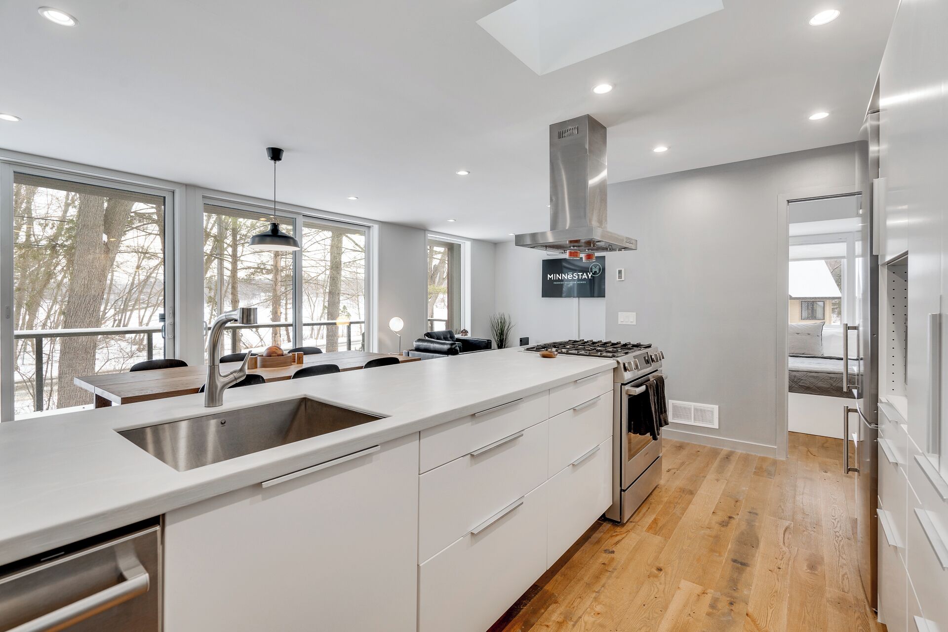 A modern kitchen with a large island, stainless steel appliances, and a view of trees through large windows.
