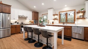 A modern kitchen with wooden cabinets, stainless steel appliances, a central island with bar stools, and a window with a view of trees.