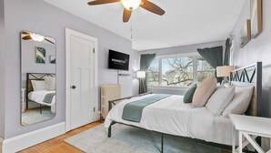 A bedroom with a bed, a ceiling fan, a TV, a window with curtains, and a radiator. - Lyndale Pleasant Getaway (Minneapolis)