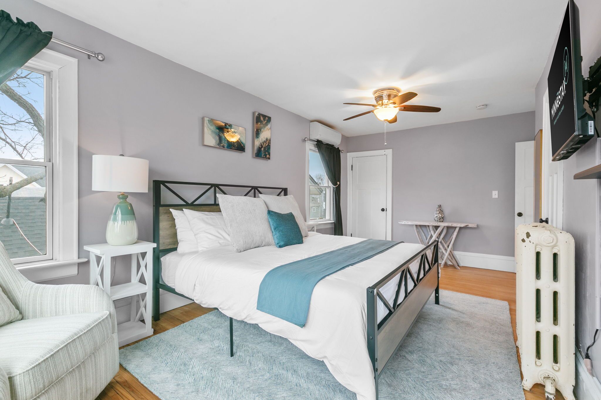A bedroom with a bed, bedside table, lamp, artwork, ceiling fan, and a radiator.