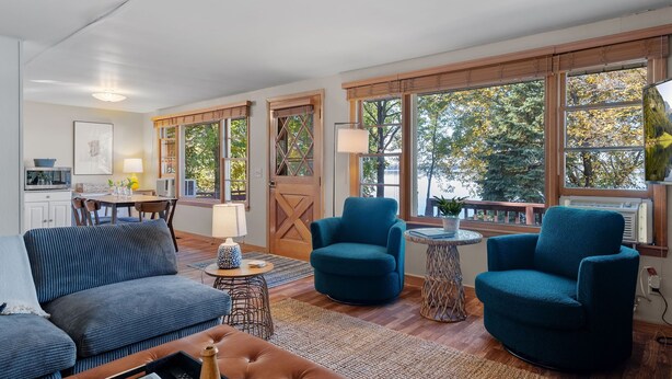 A spacious living room with a sofa, two armchairs, a coffee table, and a fireplace. There are large windows with wooden frames and a view of trees. - Lake Pulaski Getaway (Buffalo)