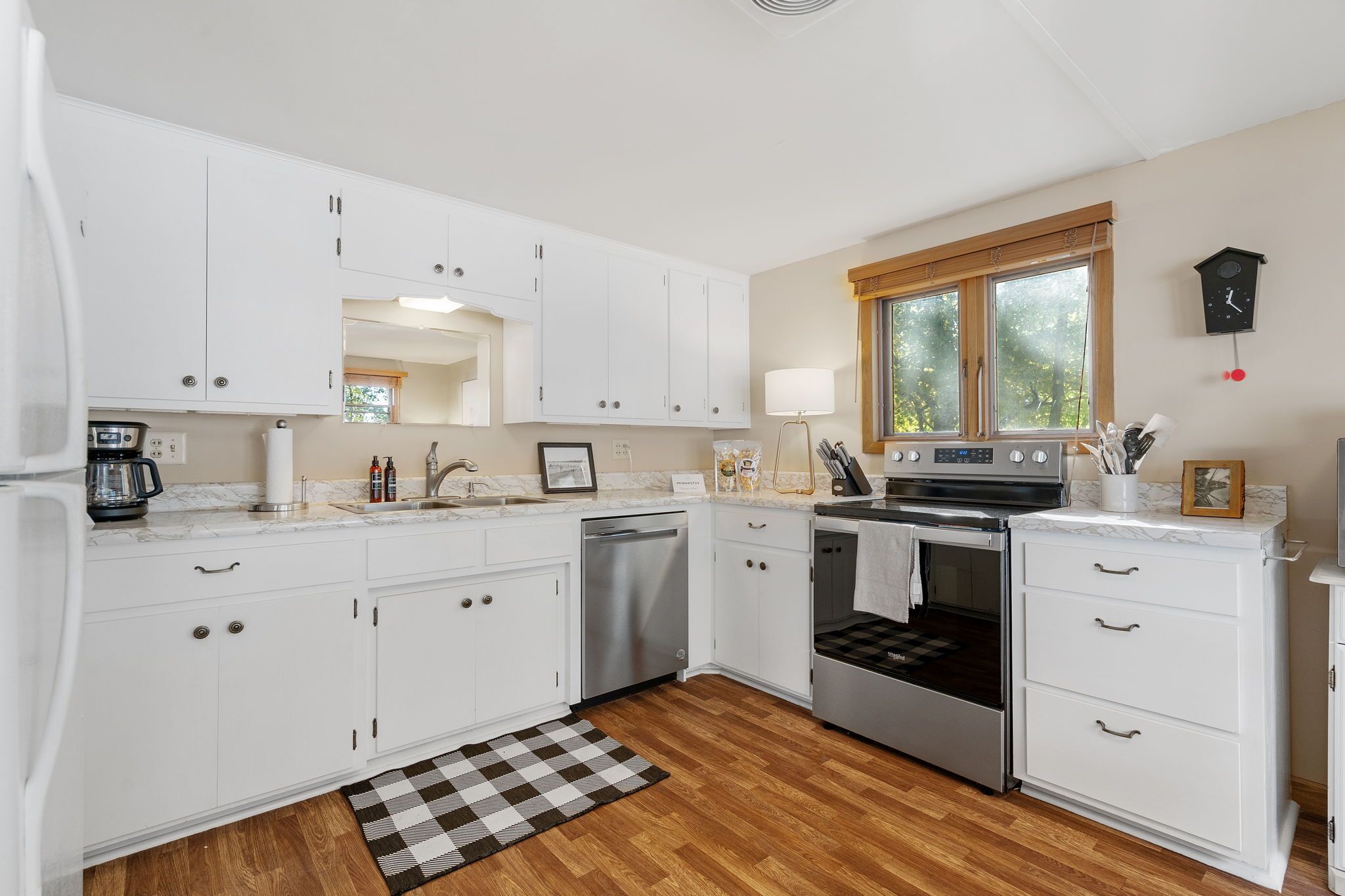 A modern kitchen with white cabinets, a dishwasher, an oven, and a window with a view of trees.