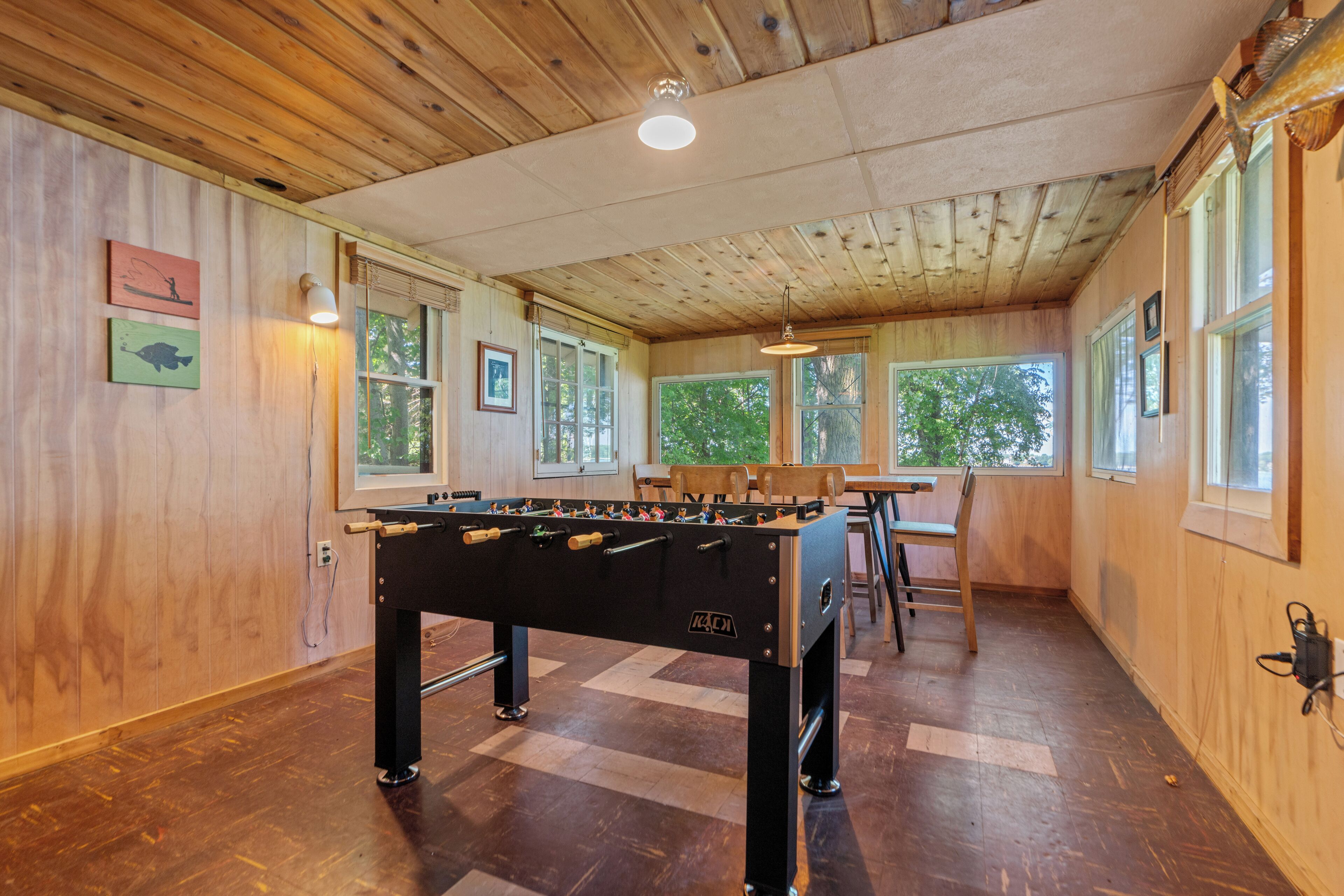 A wooden-floored room with a foosball table, framed pictures on the walls, and large windows.