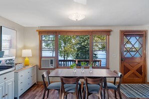 A dining area with a table and chairs, a lamp, and a view of trees and water through large windows. - Lake Pulaski Getaway (Buffalo)