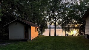 A small wooden cabin with a porch, a grassy yard, and a view of a lake at sunset. - Lake Pulaski Getaway (Buffalo)