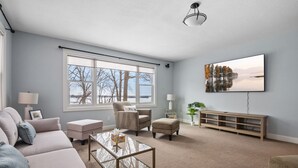 A living room with a sofa, armchairs, a coffee table, a TV, and a large window with a view of trees and a lake. - Minnetonka Dream (Wayzata)