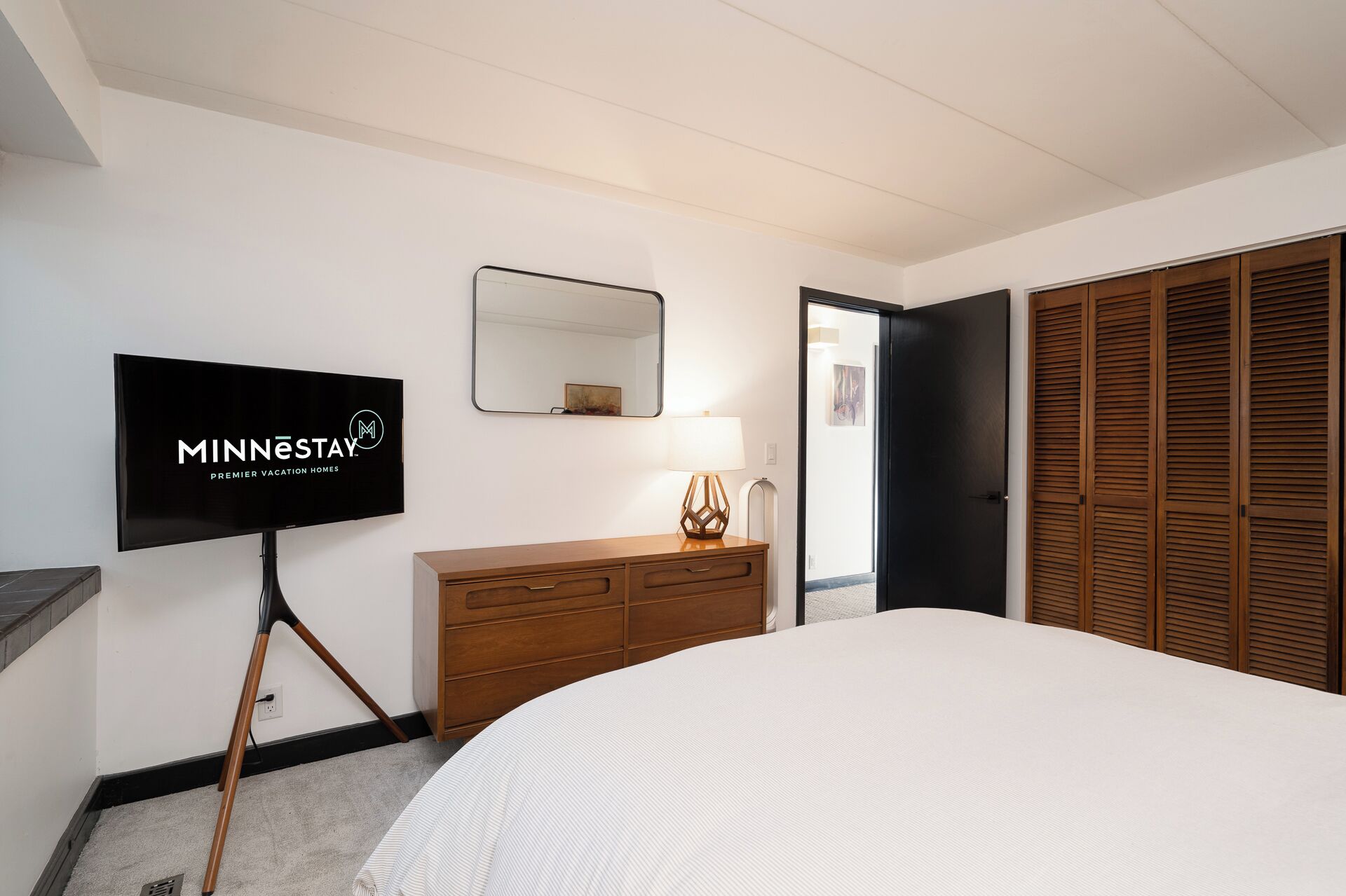 Une chambre d’hôtel avec un lit, une commode en bois, un téléviseur diffusant une émission sur les « meilleures locations de vacances du Minnesota », un miroir et une porte donnant sur un placard.