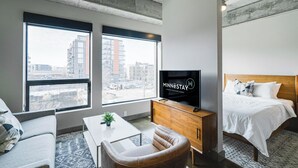 A modern hotel room with a large window, a flat-screen TV, a bed, a sofa, and a wooden cabinet. - Sable 302 - Alcove (Minneapolis)