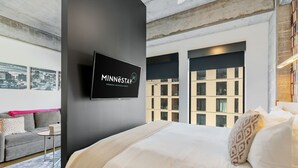 A modern hotel room with a large bed, a flat-screen TV mounted on the wall, a gray sofa, and large windows offering a city view. - Sable 304 - Alcove (Minneapolis)