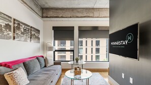 A modern living room with a gray sofa, a glass coffee table, and a cityscape view through large windows. - Sable 304 - Alcove (Minneapolis)