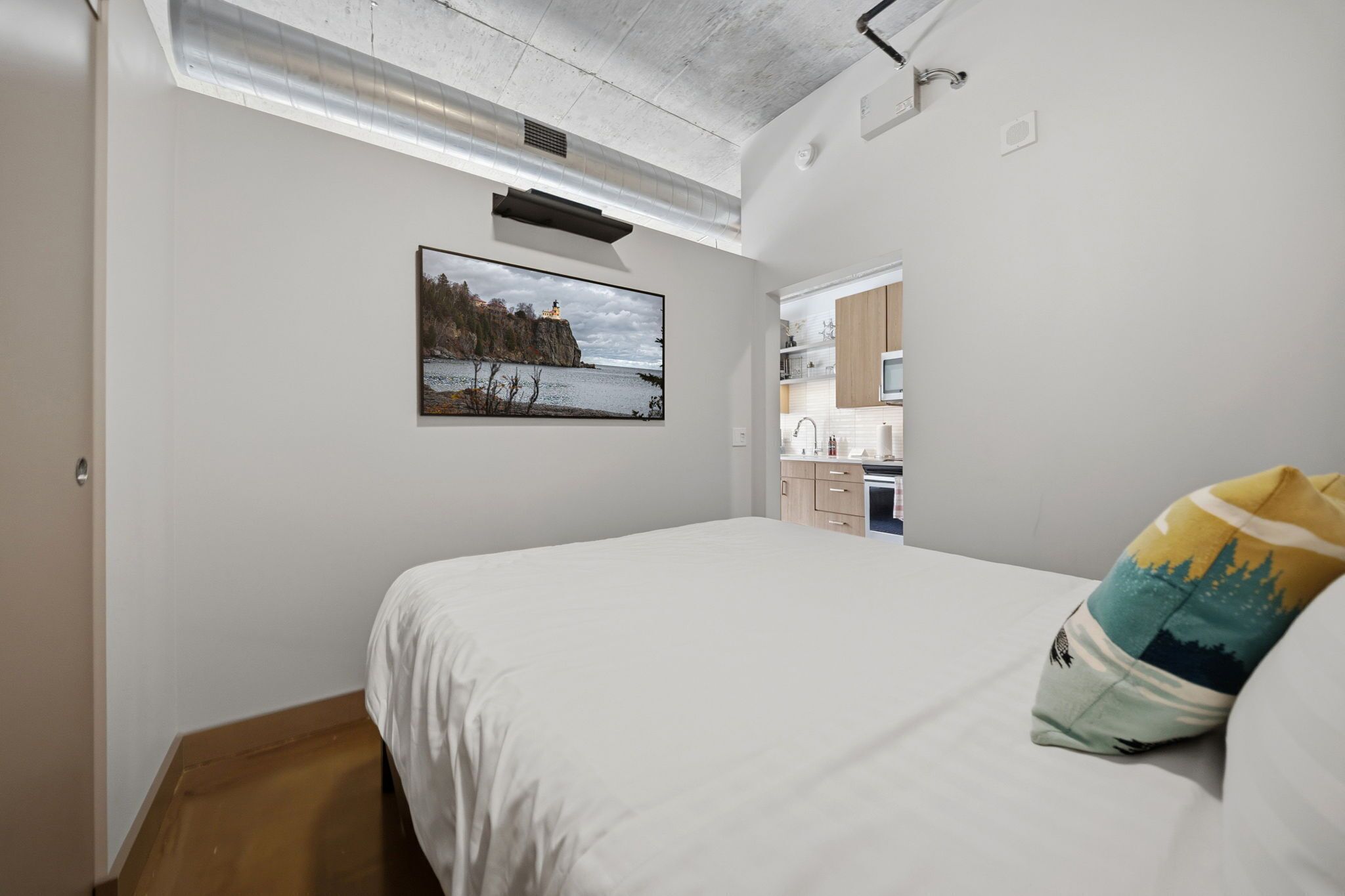 A bedroom with a bed, a pillow, a picture on the wall, and a view into the kitchen.
