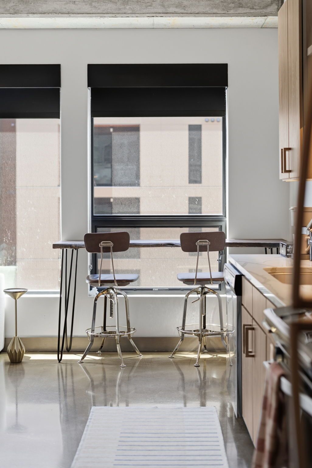 A modern kitchen with two bar stools, a window with blinds, and a view of a building outside.
