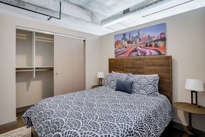 A bedroom with a bed, a nightstand, a lamp, a painting of a cityscape, and a wardrobe. - Sable 207 - One Bedroom (Minneapolis)