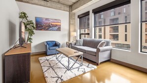 A modern living room with a sofa, a coffee table, and a TV on a wooden cabinet. There is a painting on the wall and a window with a view of buildings. - Sable 204 - One Bedroom (Minneapolis)